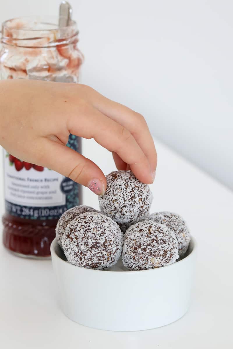 A hand reaching for a coconut 'Lamington' bliss ball in a bowl.