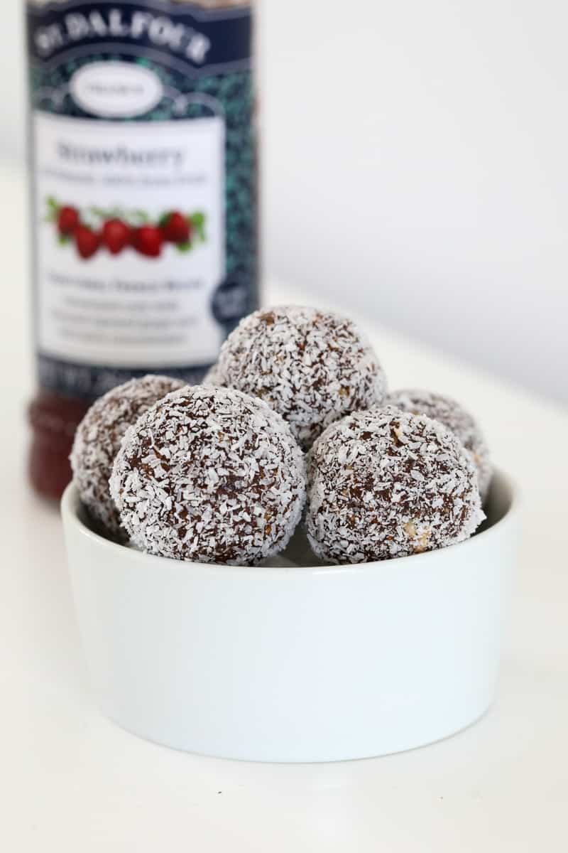 A bowl of date and almond  protein balls coated in coconut, in front of a jar of strawberry jam.