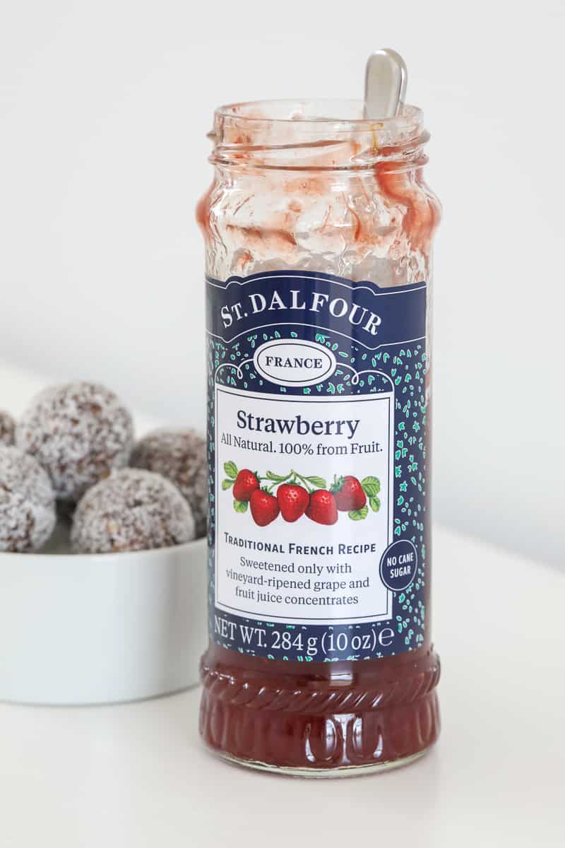 A jar of strawberry fruit spread in front of a bowl of bliss balls.