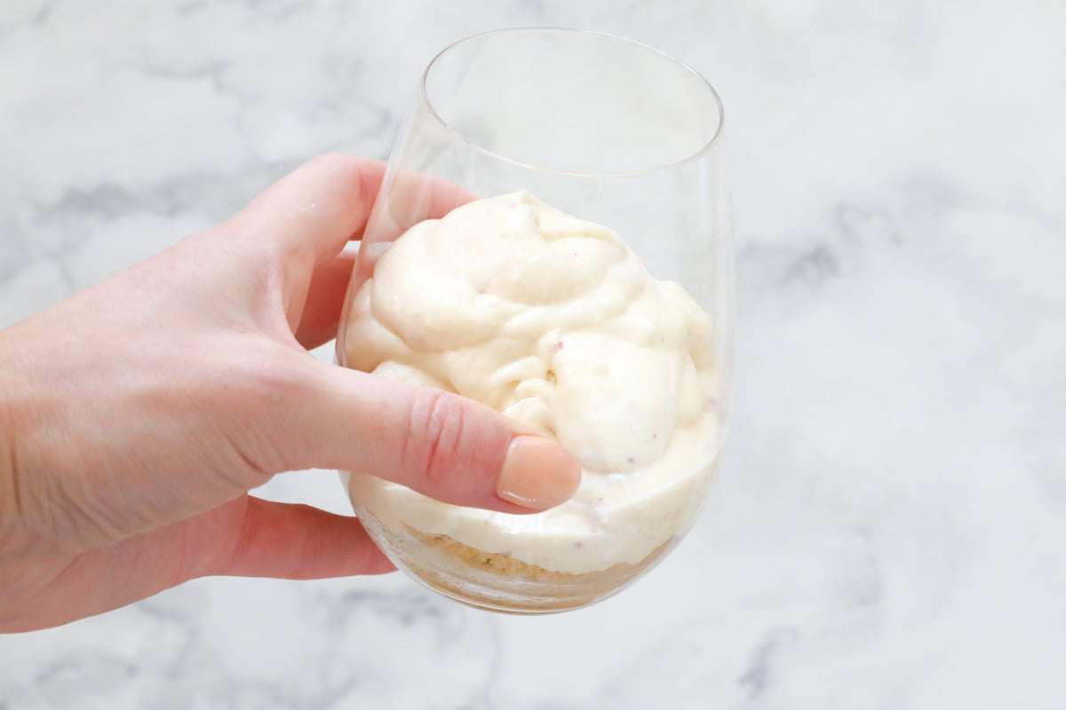 A hand holding a stemless wine glass with a biscuit crumb base and creamy cheesecake filling