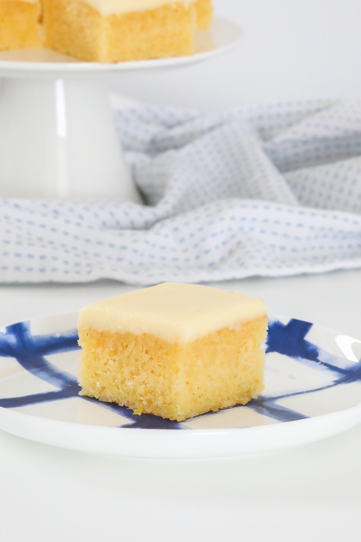 A piece of baked lemon slice on a blue and white plate, and a cake stand blurred in the background.