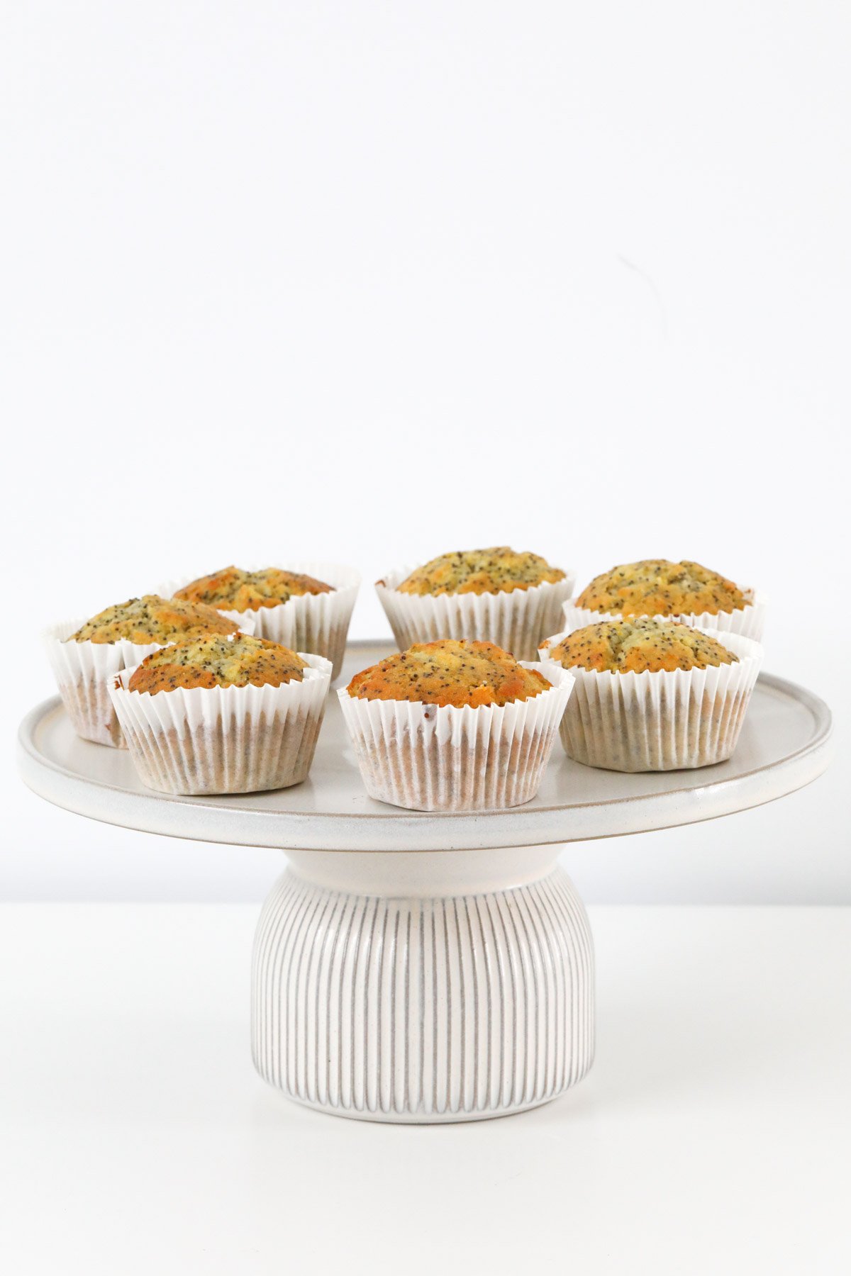 A chunky-based white cake stand with six orange & poppyseed muffins arranged on it