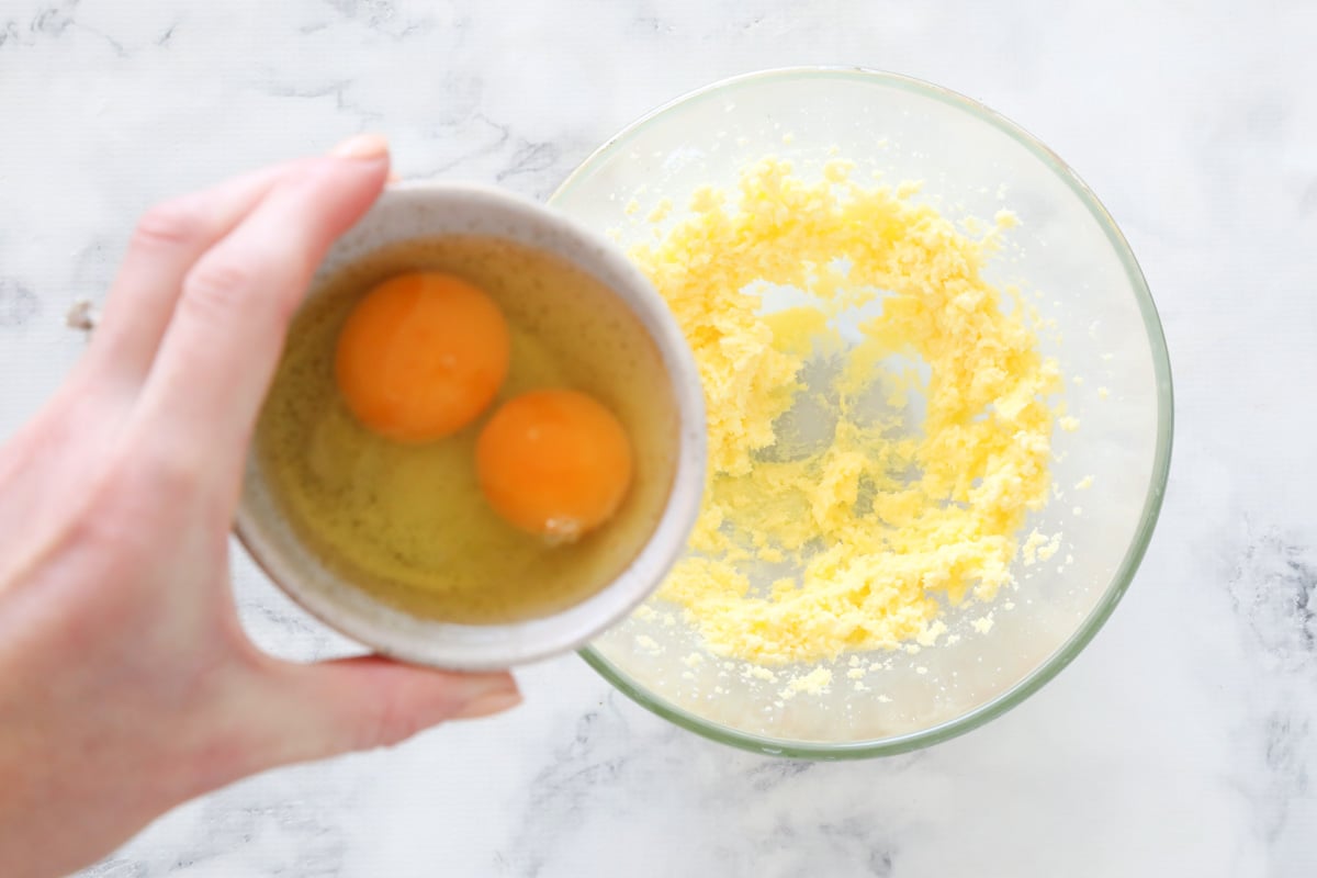 Two cracked eggs in a ramekin being added to a glass bowl with creamed butter and sugar
