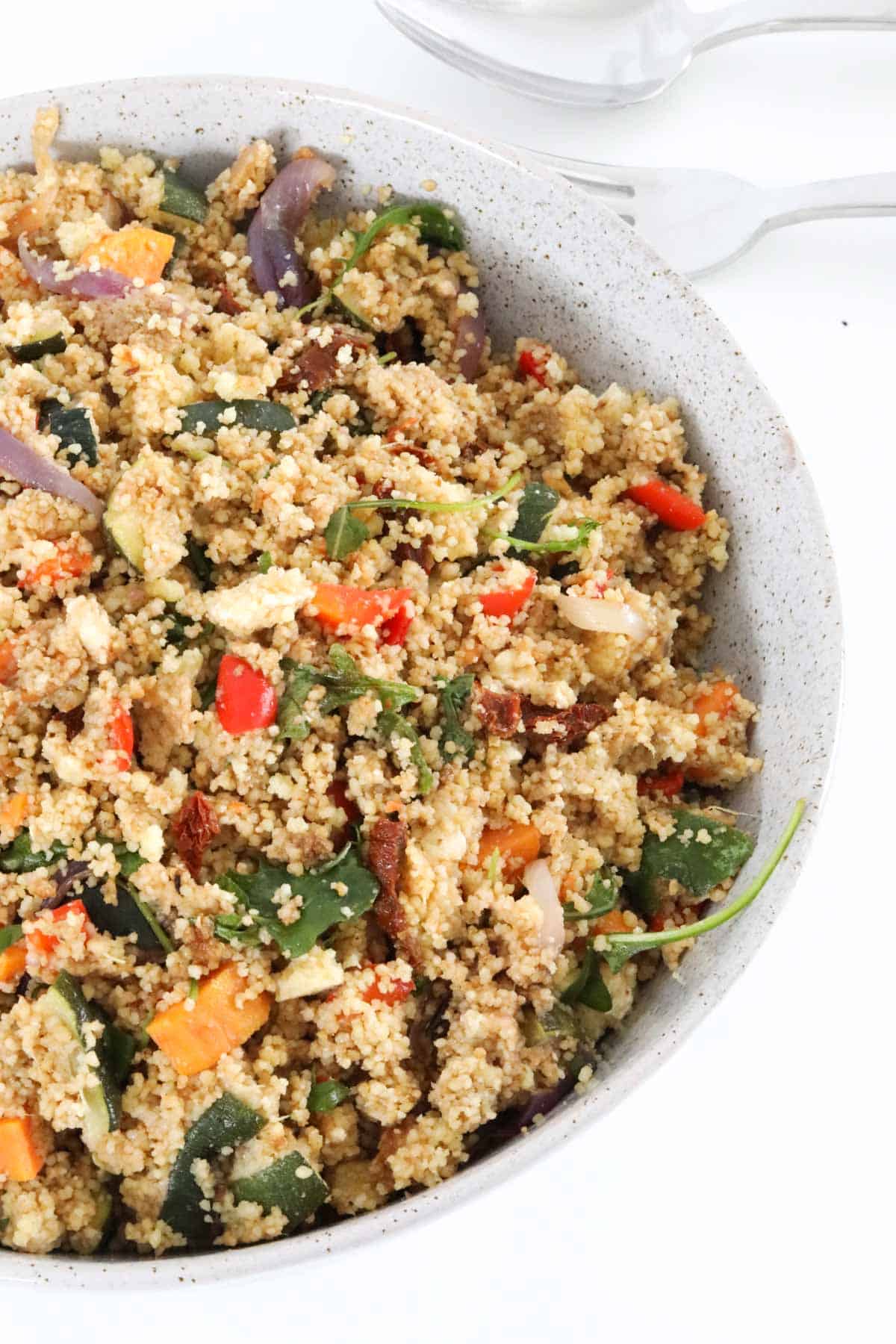 A close up image of roasted vegetable and couscous salad in a speckled bowl with salad servers just visible on the bench in the background