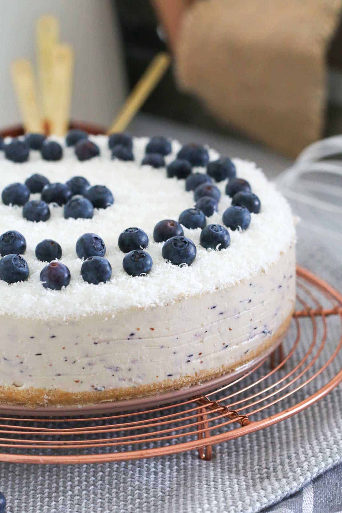 A blueberry cheesecake on a wire stand, topped with fresh blueberries and grated white chocolate.