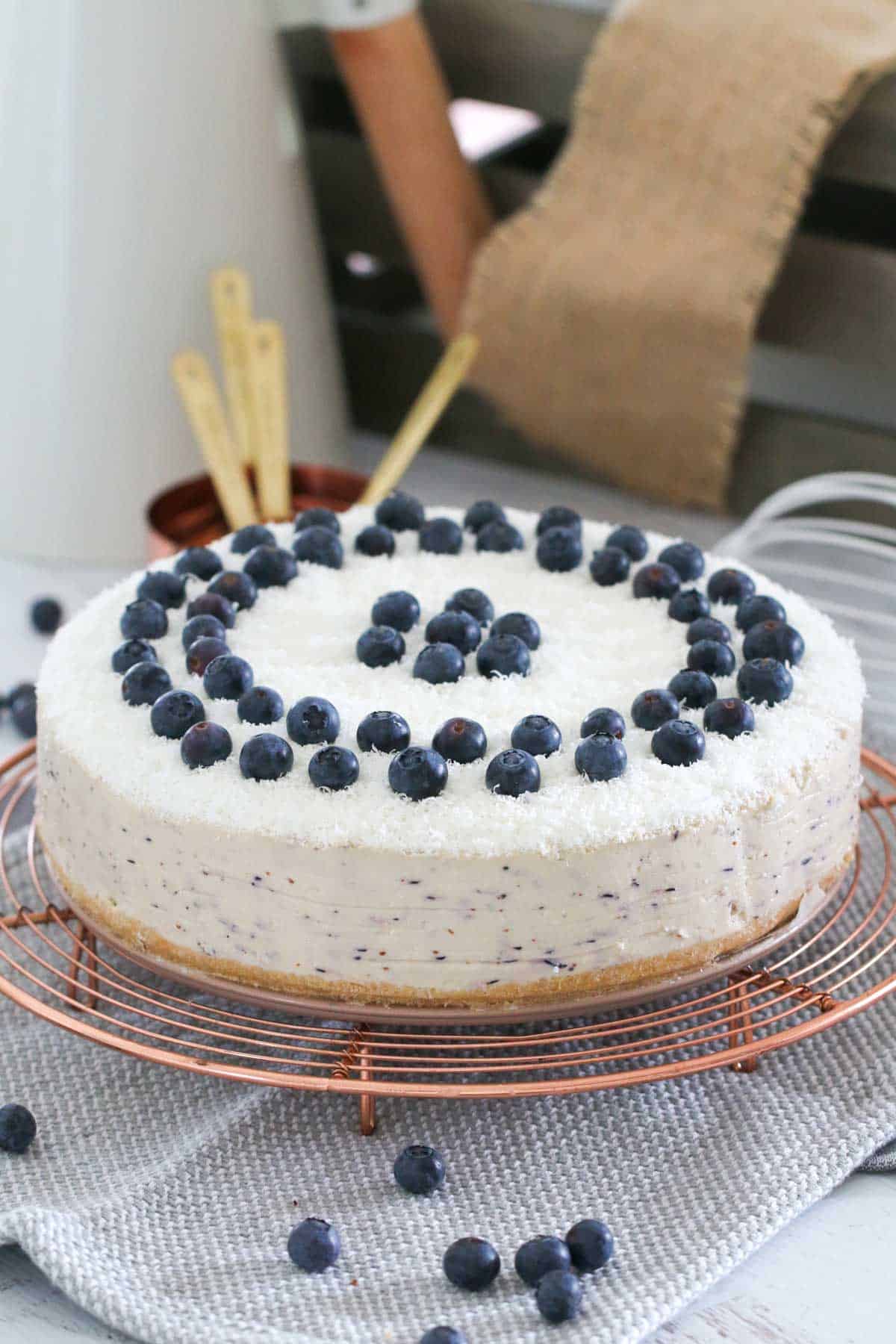 A white cheesecake on a wire cake stand topped with blueberries.
