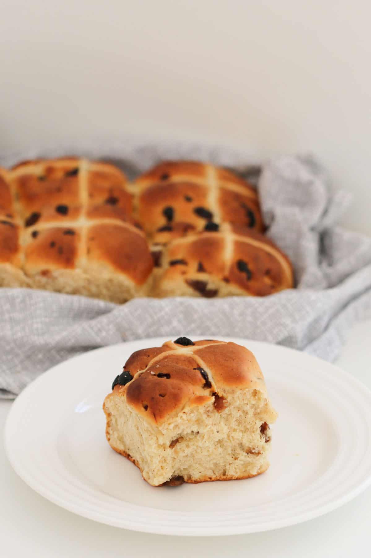 A hot cross bun on a white plate with a batch of buns in the background.