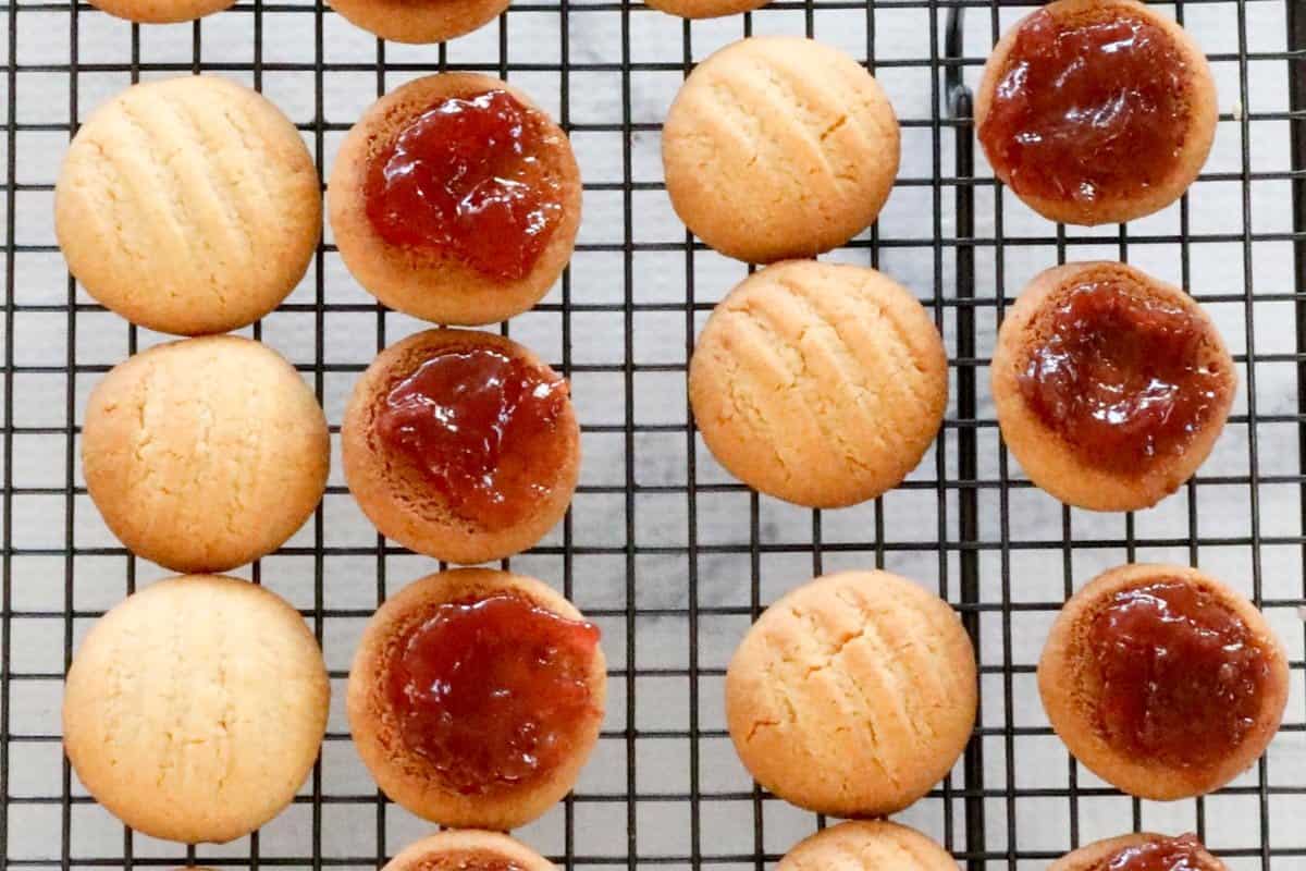 A batch of coconut cookies on a wire rack, with half spread with strawberry jam