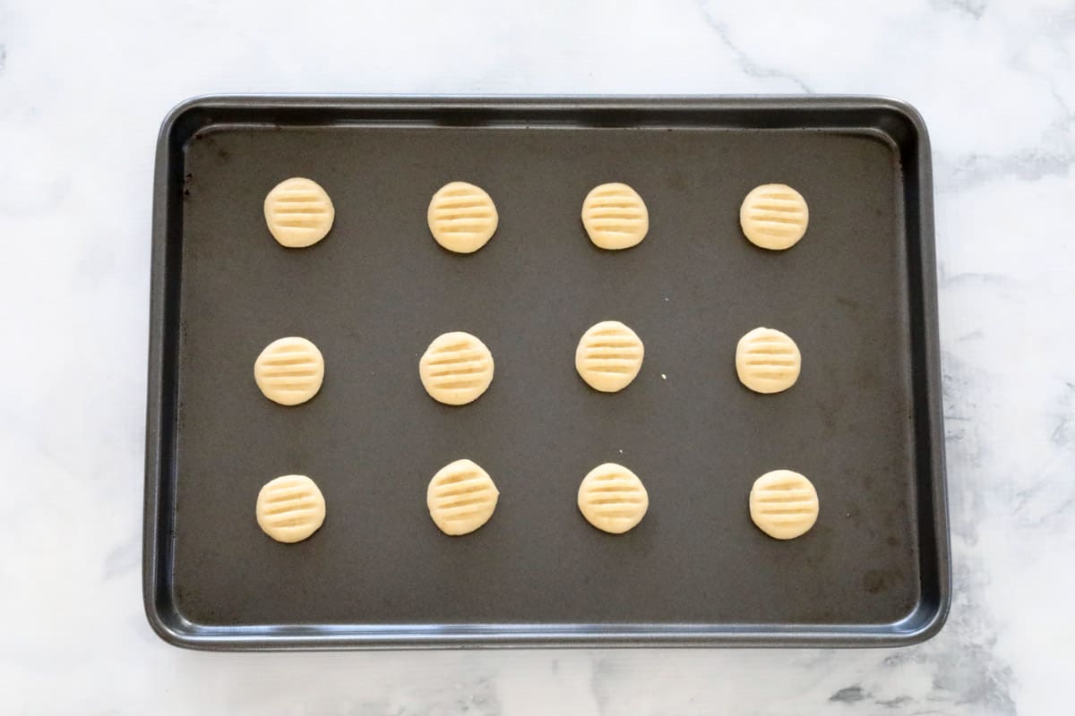 12 biscuits pressed down with a fork on a rectangular baking tray on a marble counter top