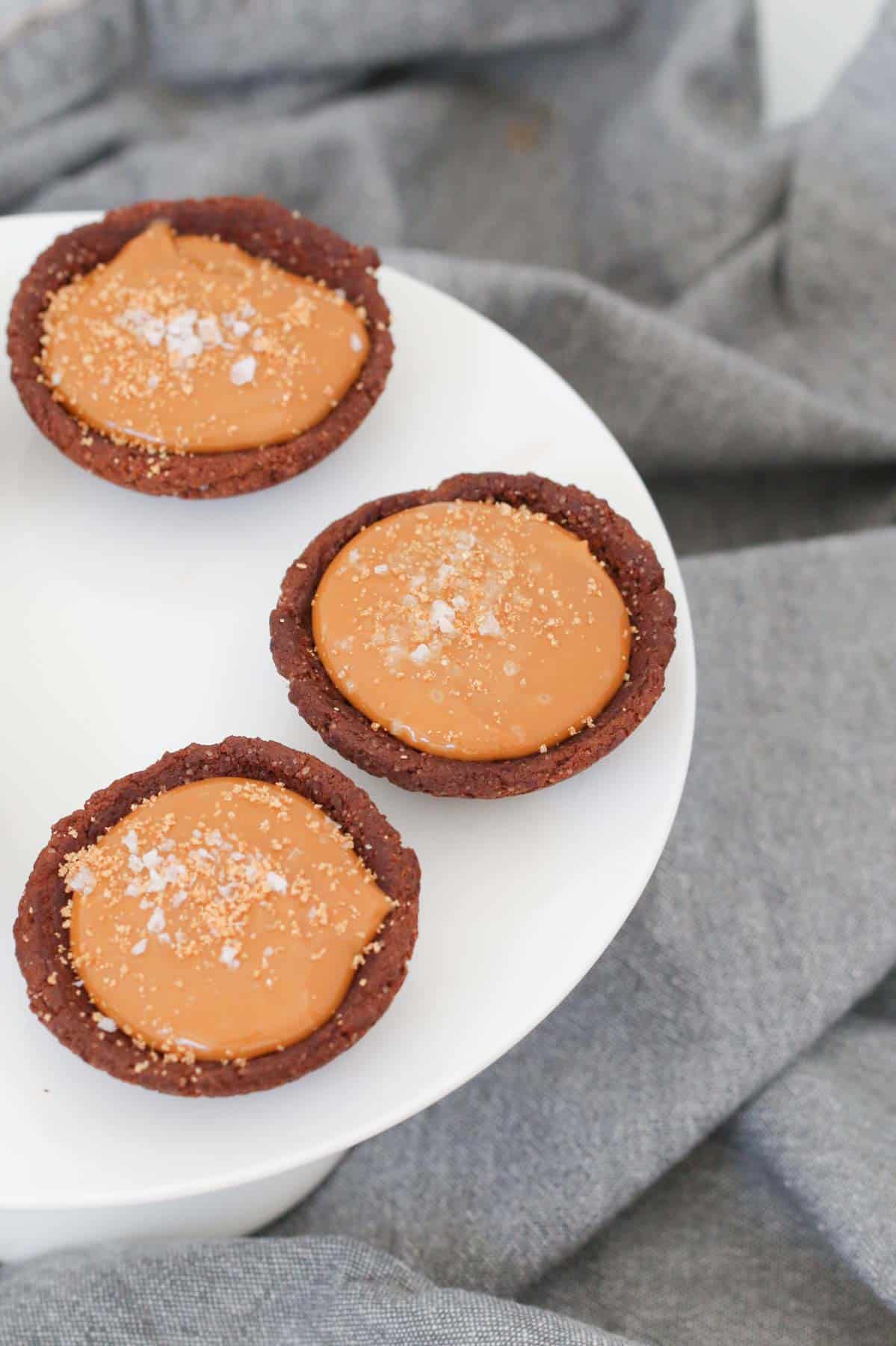 Three mini caramel tarts in chocolate cookie shells on a white cake stand.