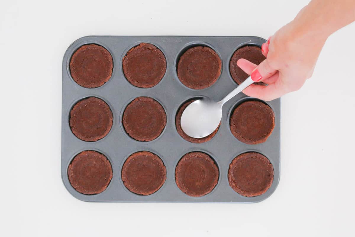 A spoon pressing down chocolate biscuits into a patty pan tin.