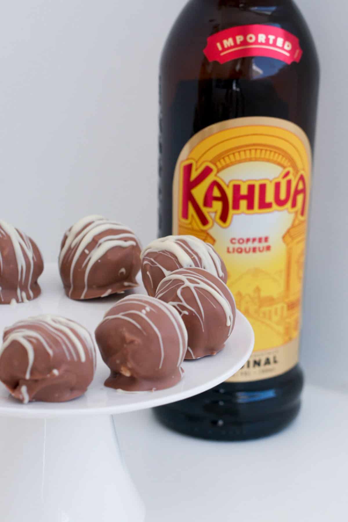 A white cake platter with Cheesecake balls in the foreground and a bottle of kahlua in the background.