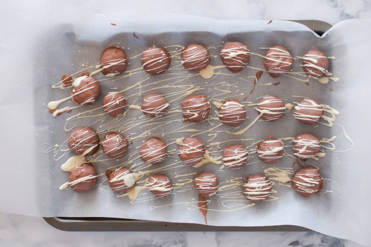 A tray lined with baking paper and chocolate coated cheesecake balls on top, with drizzled white chocolate.