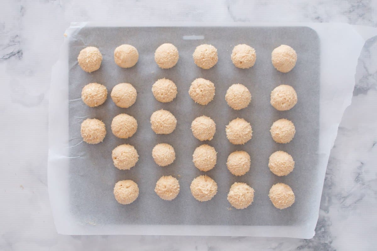 A baking tray lined with baking paper and rolled cheesecake balls on it.