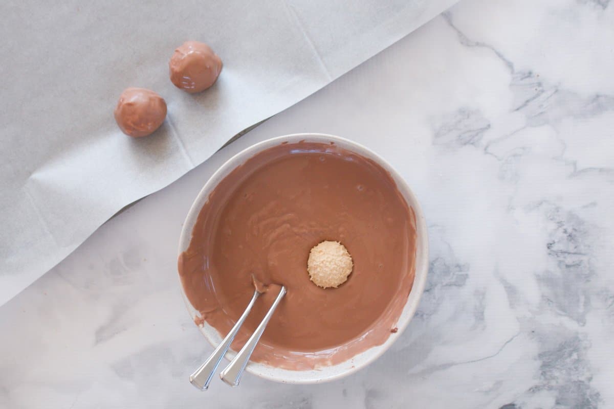 A bowl with melted chocolate, a ball and two forks, and a piece of baking paper with two chocolate covered balls on it.