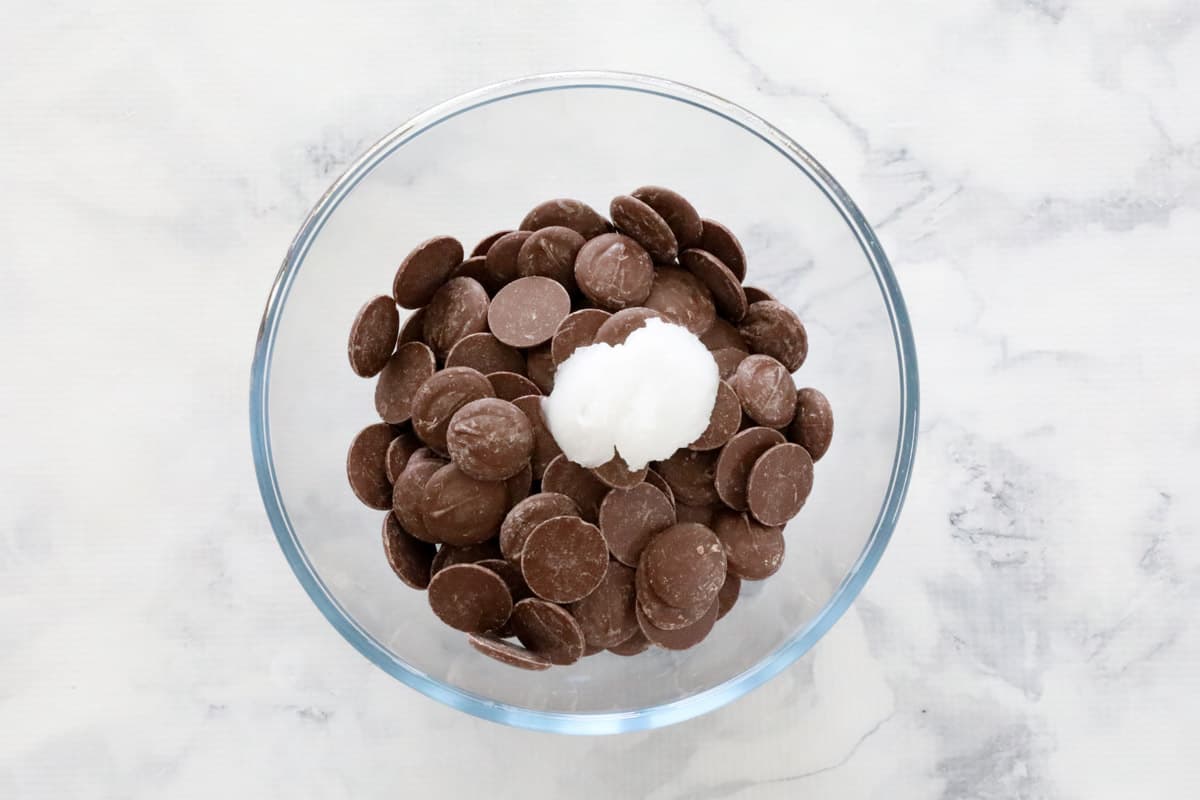 A glass bowl on a marble counter with chocolate melts and coconut oil