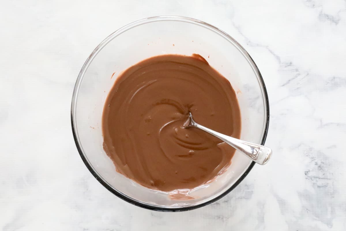 Melted milk chocolate with a silver spoon resting against the side of a glass bowl on a marble counter