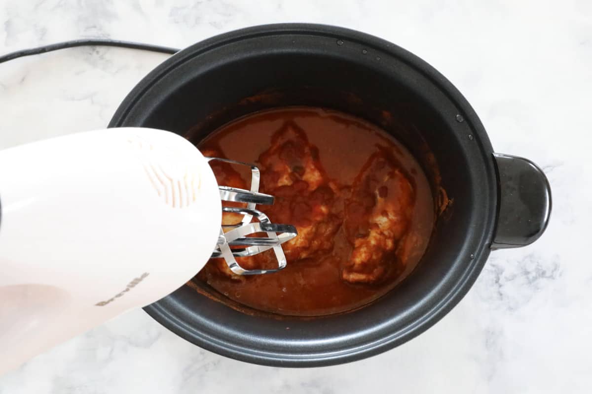 Chicken being shredded by hand held beaters in a slow cooker pot