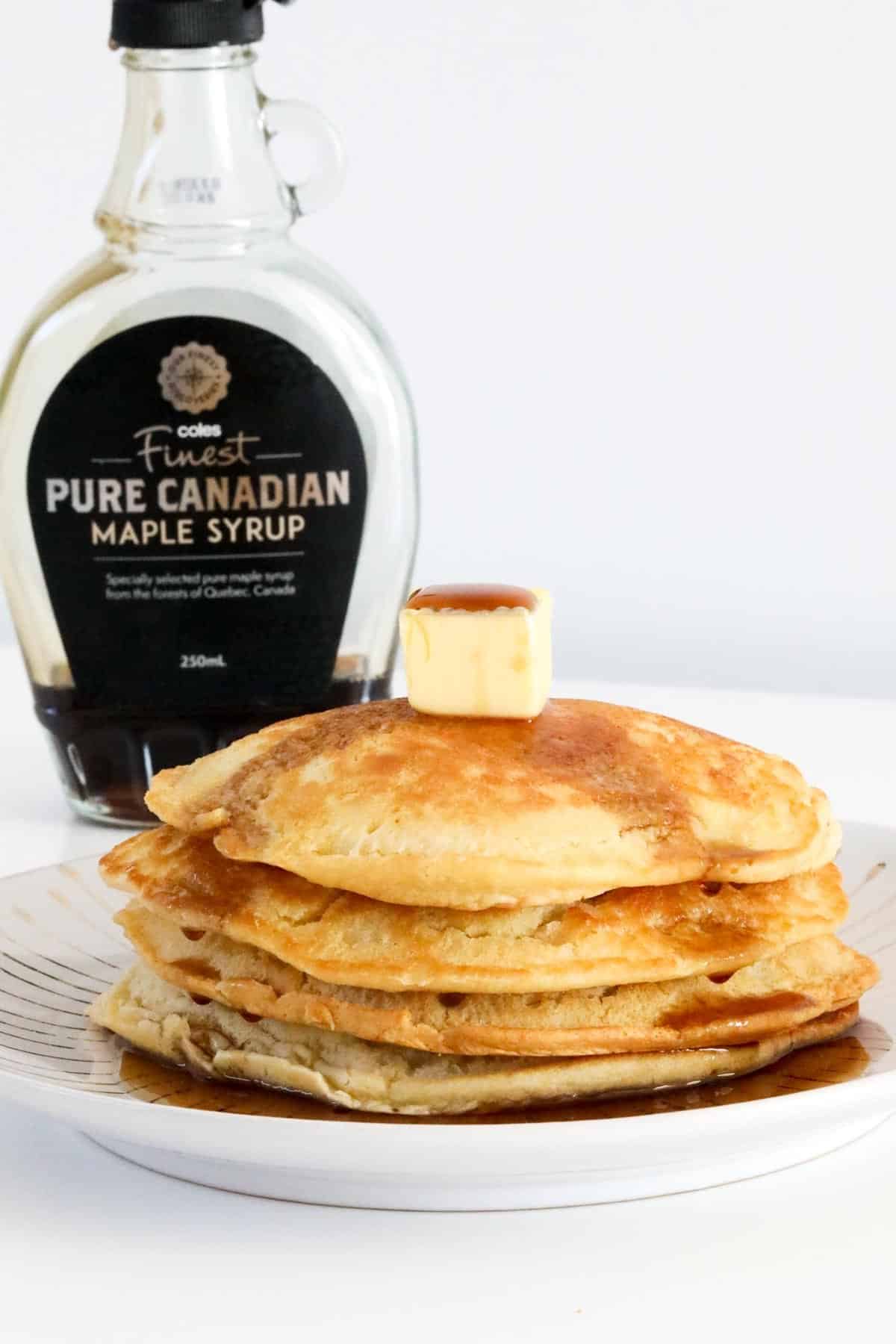 A jar of maple syrup behind a stack of pancakes.
