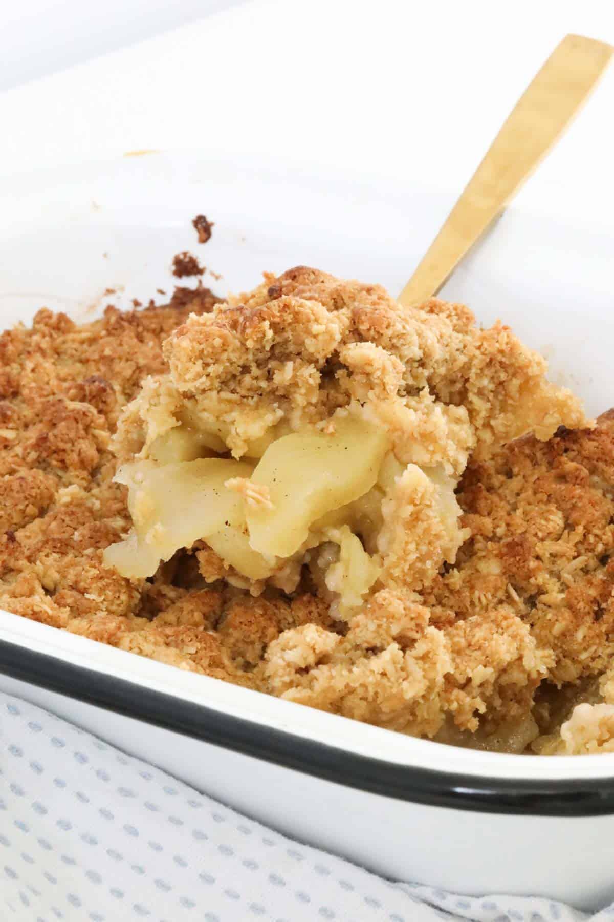 A gold serving spoon lifting out a serving of Apple Crumble from the enamel baking dish