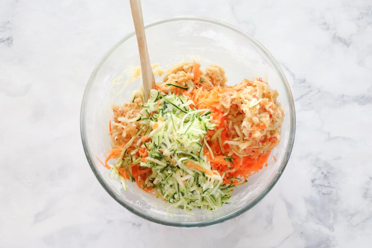 Grated carrot, zucchini and apple, added to mixture in glass bowl.