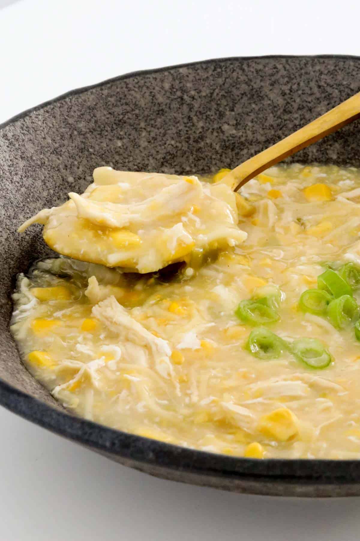 A gold spoon in a bowl of chicken and corn soup, sprinkled with sliced spring onions.