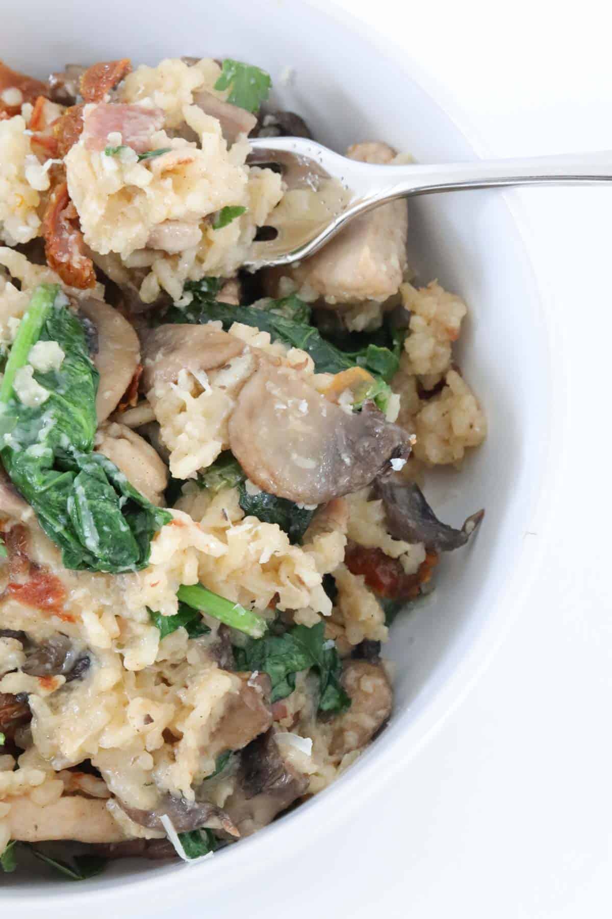 A close up of chicken risotto with mushrooms and spinach in a white bowl with a fork