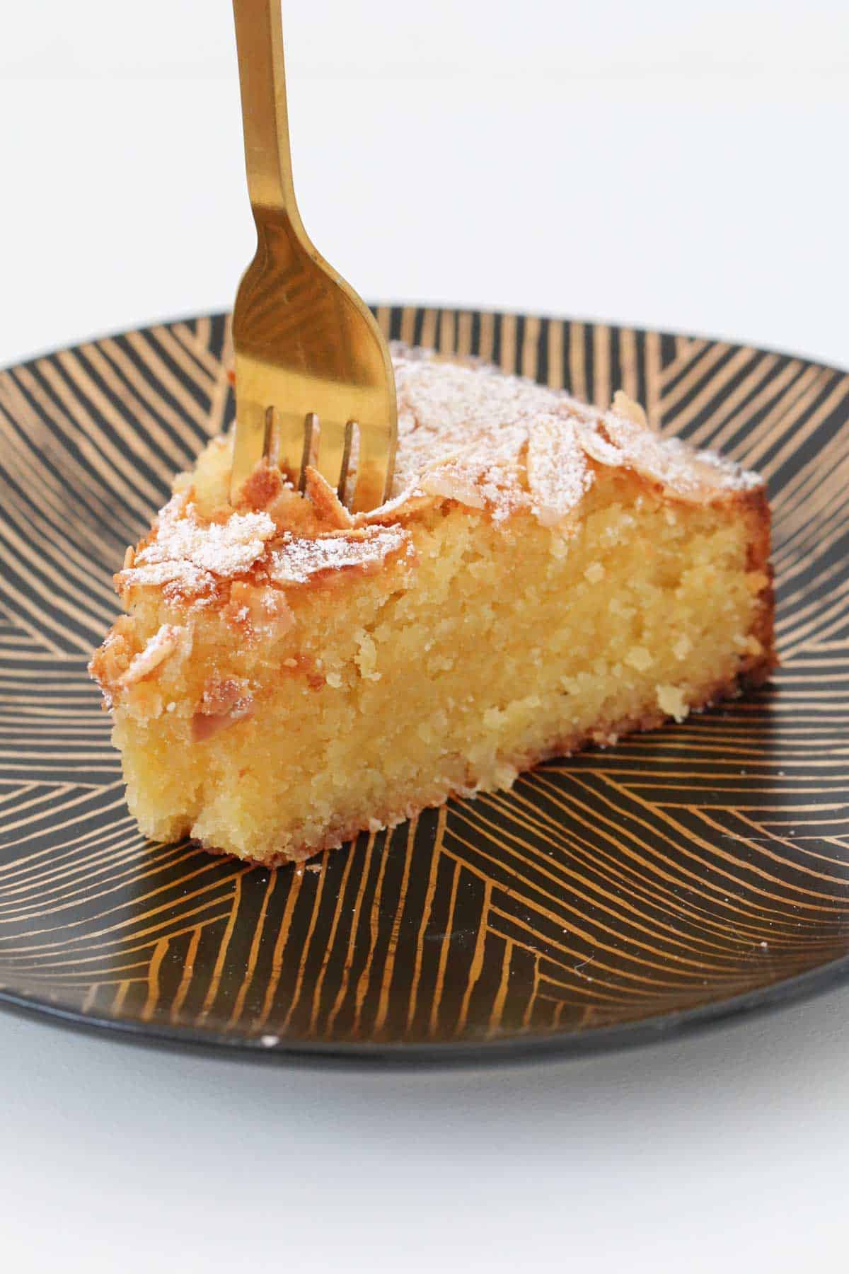 A coconut & almond cake served on a black plate with a gold fork standing up in it.