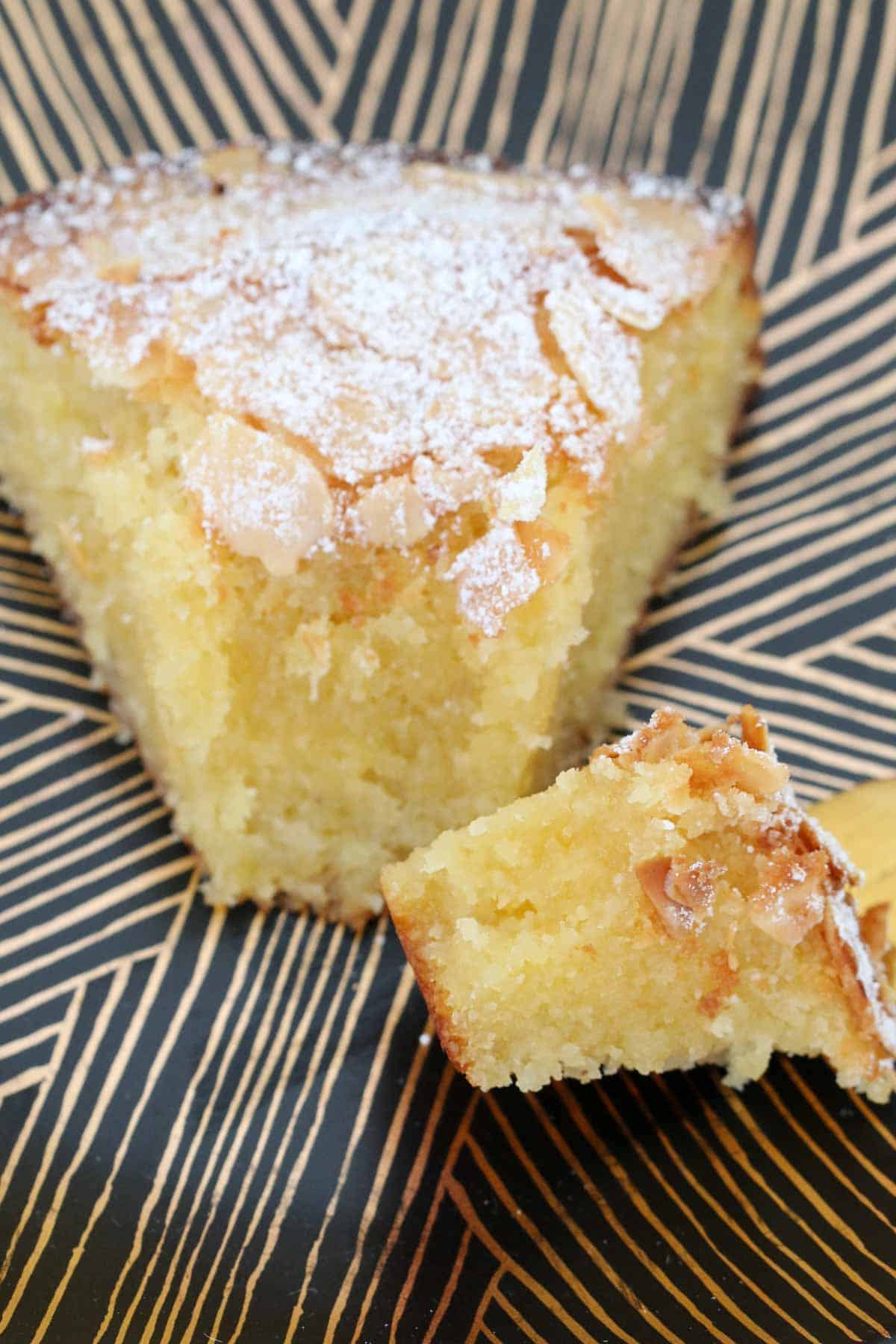 A close up of a serve of moist almond cake dusted with icing sugar, with a forkful on the side.