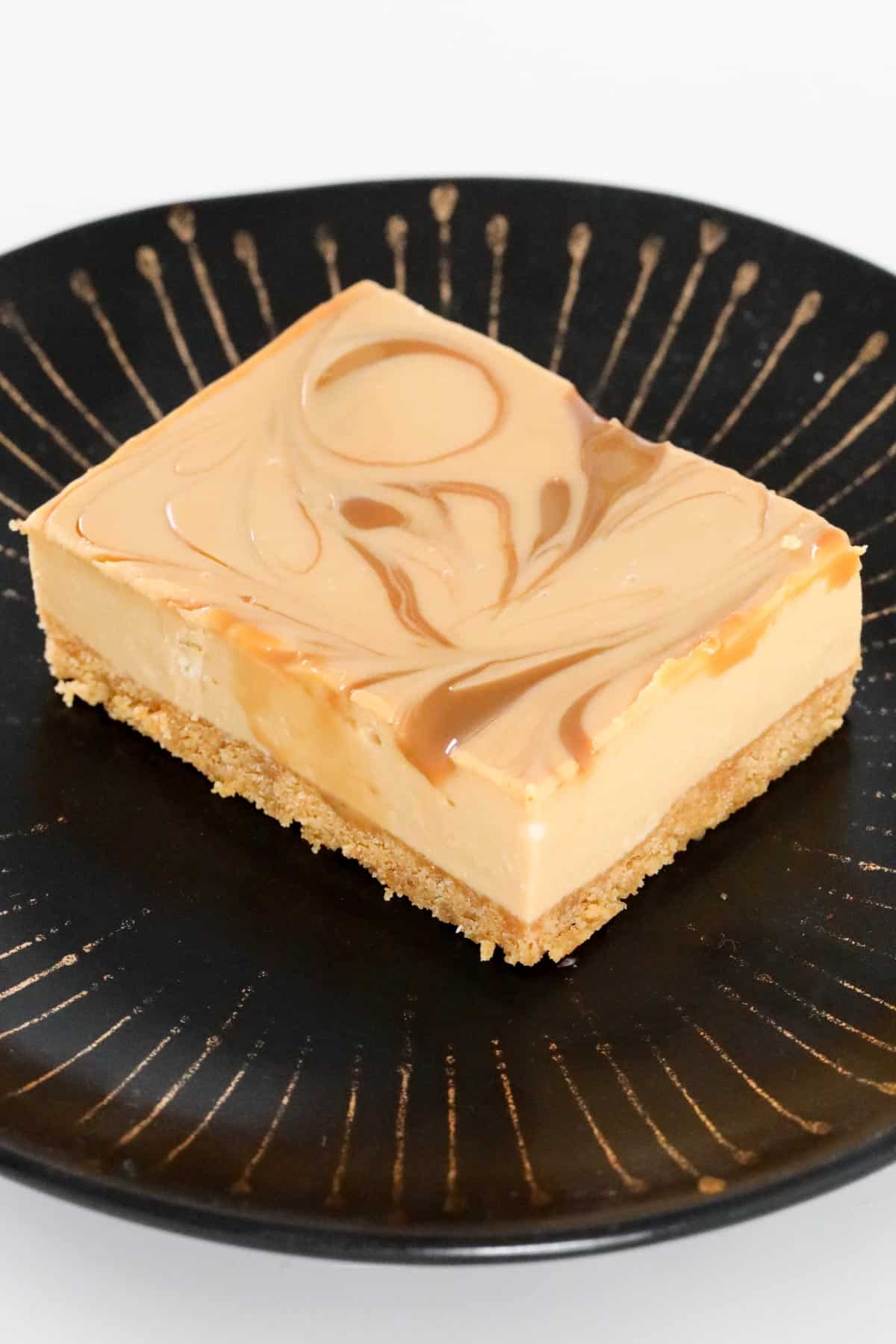 A piece of caramel cheesecake on a black and gold plate.