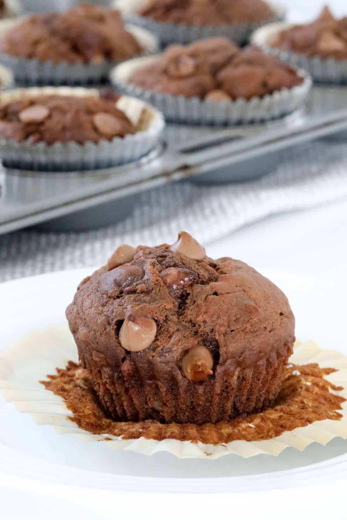 A chocolate muffin with the mffin case peeled open, in front of a tray full of baked muffins.
