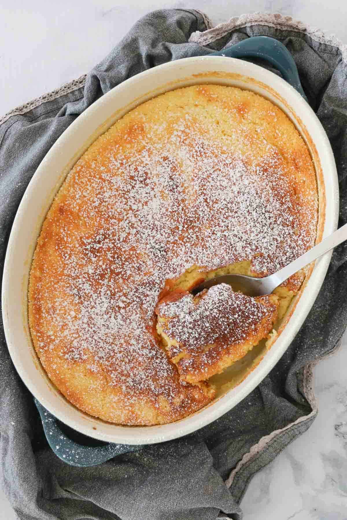 A serving spoon scooping lemon delicious pudding from an oval baking dish.