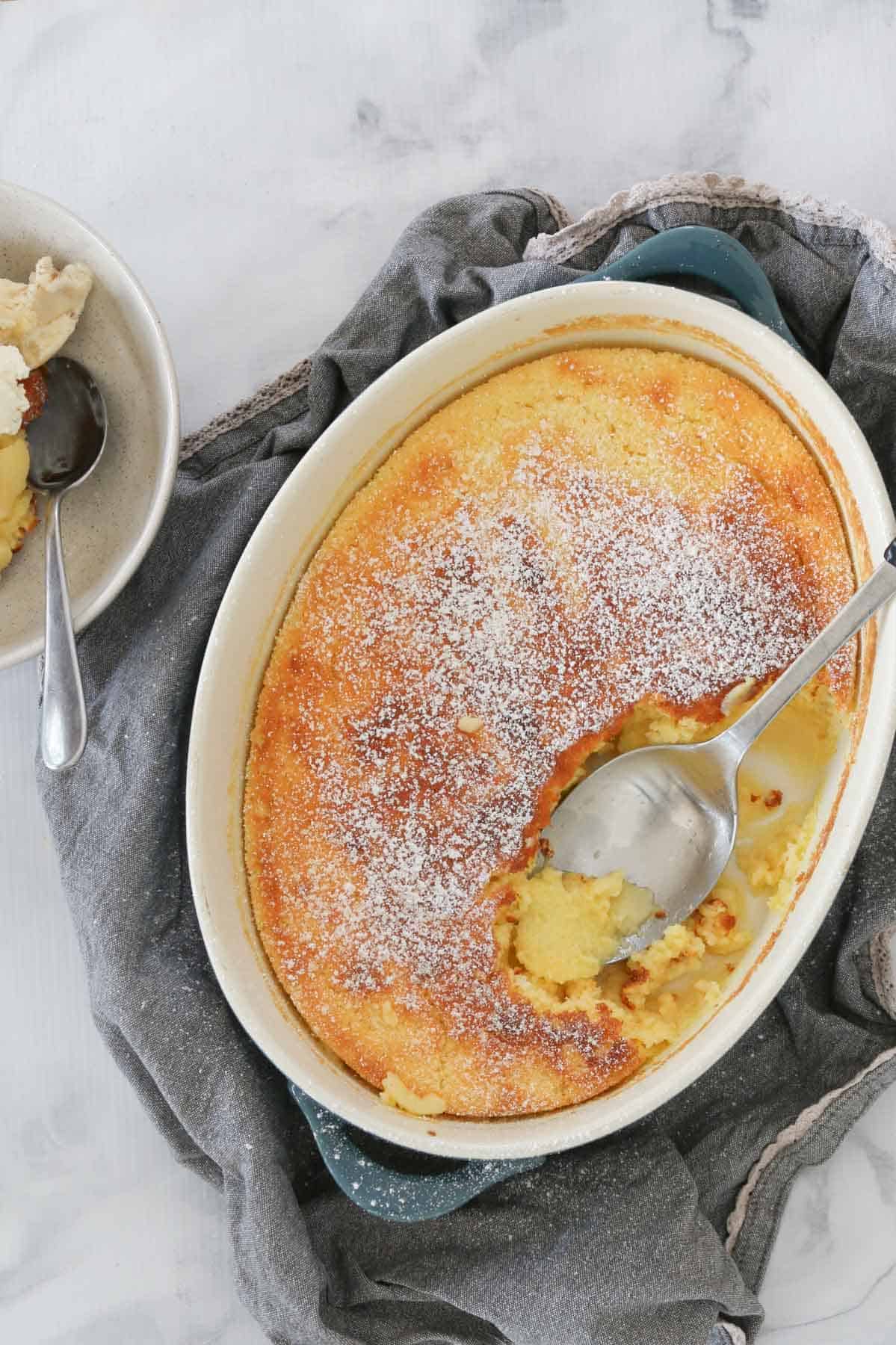 An overhead view of a white oval ovenproof baking dish filled a golden baked pudding dusted with icing sugar.