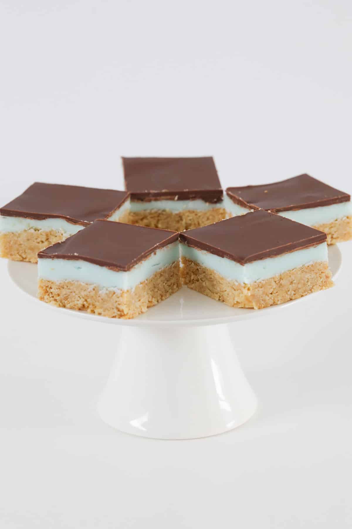 Five squares of layered peppermint slice on a white cake stand.