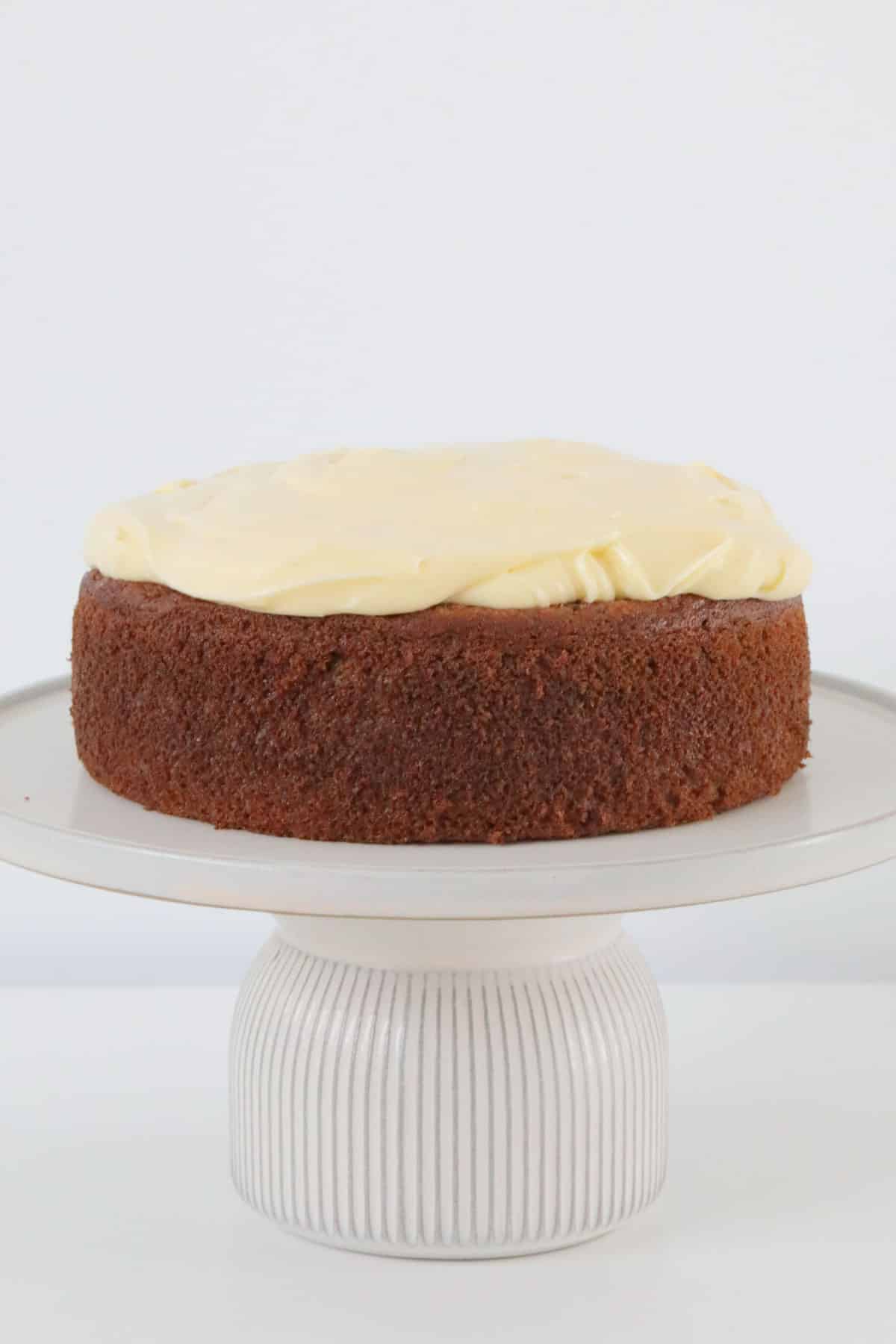 A golden brown Thermomix banana cake with thick cream cheese frosting on a white cake stand