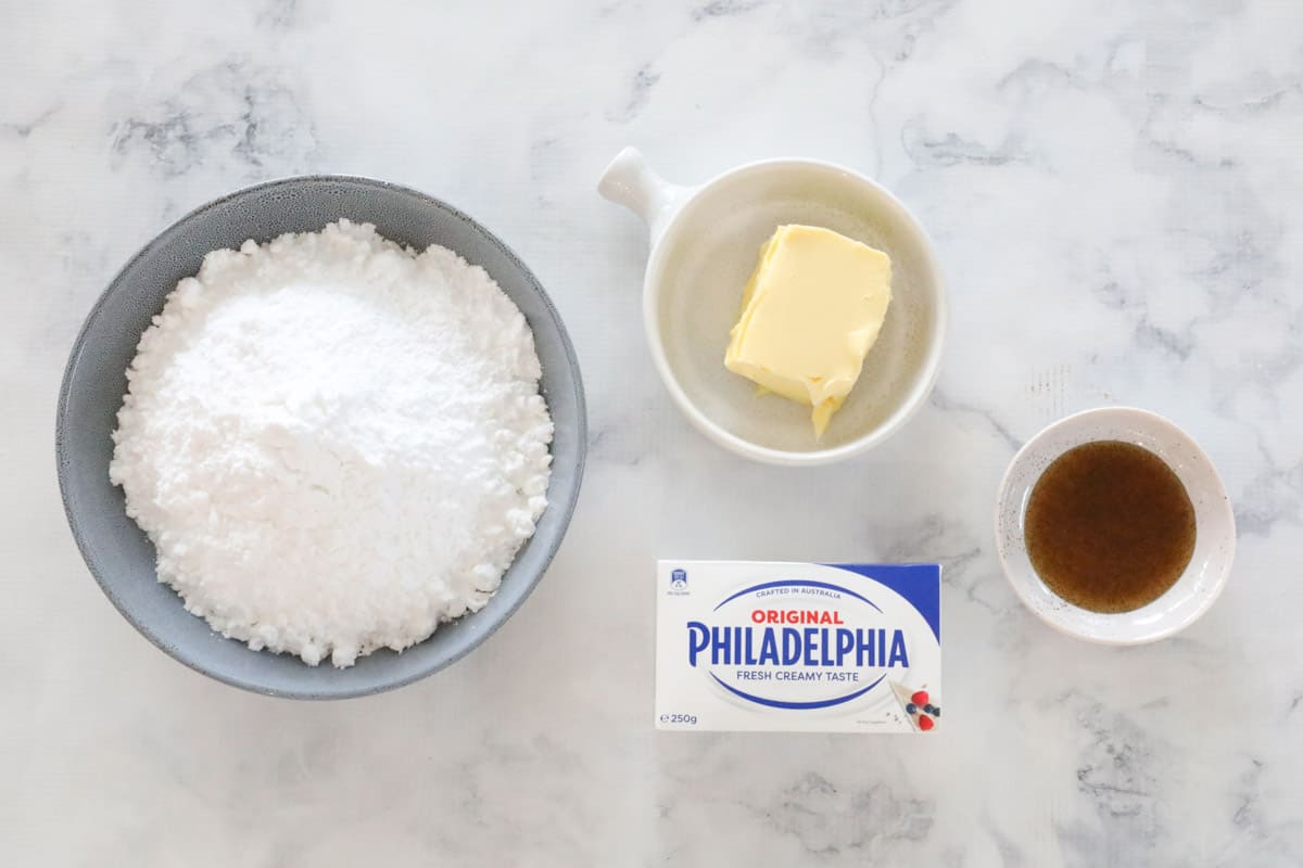 A bowl of icing sugar, ramekins with butter and vanilla extract and a block of cream cheese on a counter top