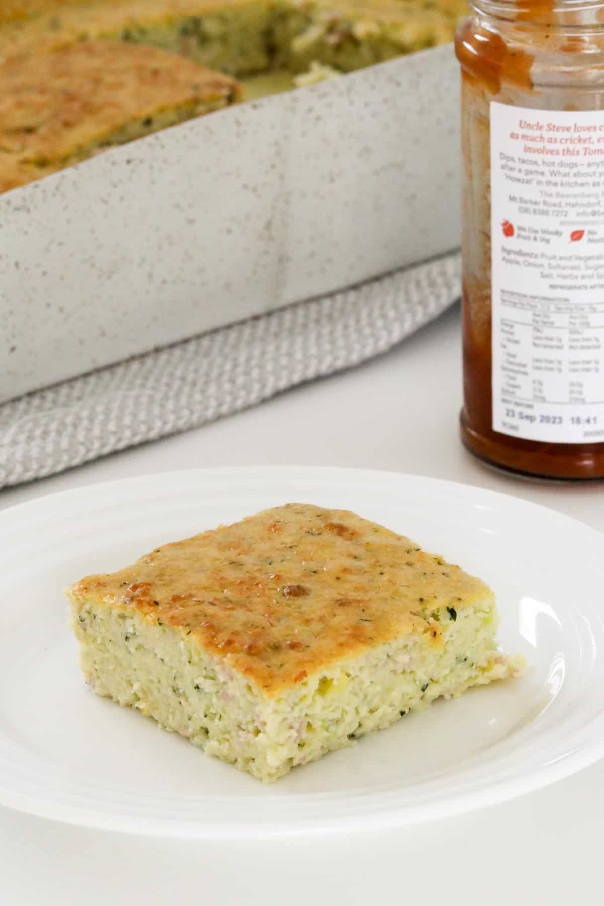 A square of savoury zucchini slice on a white plate, with a jar of chutney in the background.