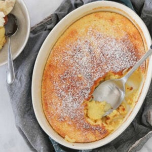 A white baking dish filled with a golden baked lemon pudding with a spoon showing the sauce beneath.