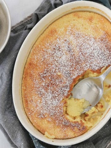 A white baking dish filled with a golden baked lemon pudding with a spoon showing the sauce beneath.