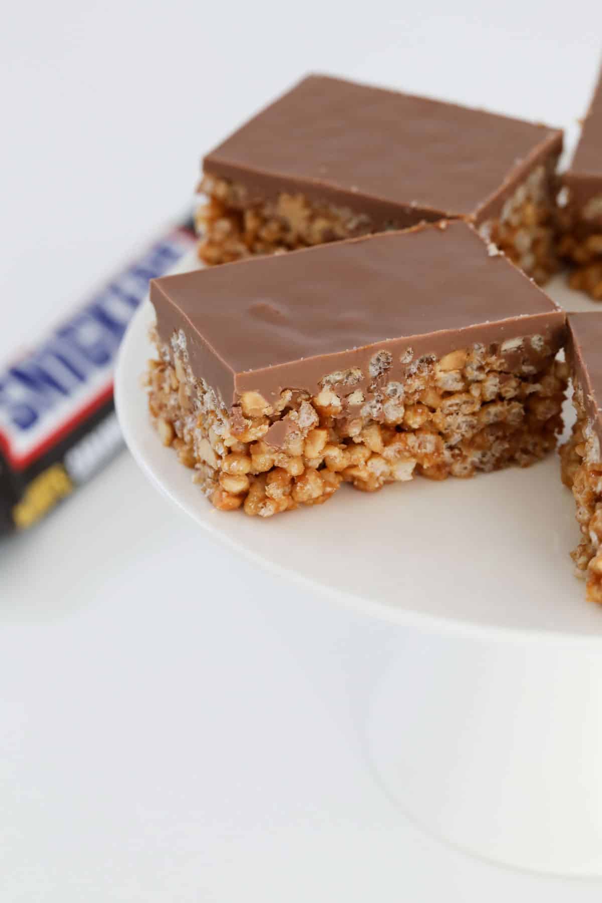 snickers slice on a white stand, topped with chocolate.
