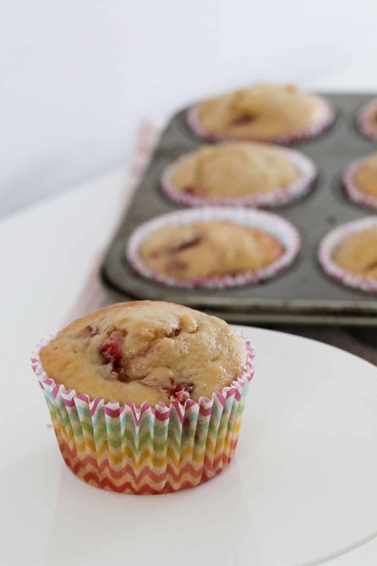 A single strawberry muffin in a colourful case on a white cake stand, with a muffin tray in the background.