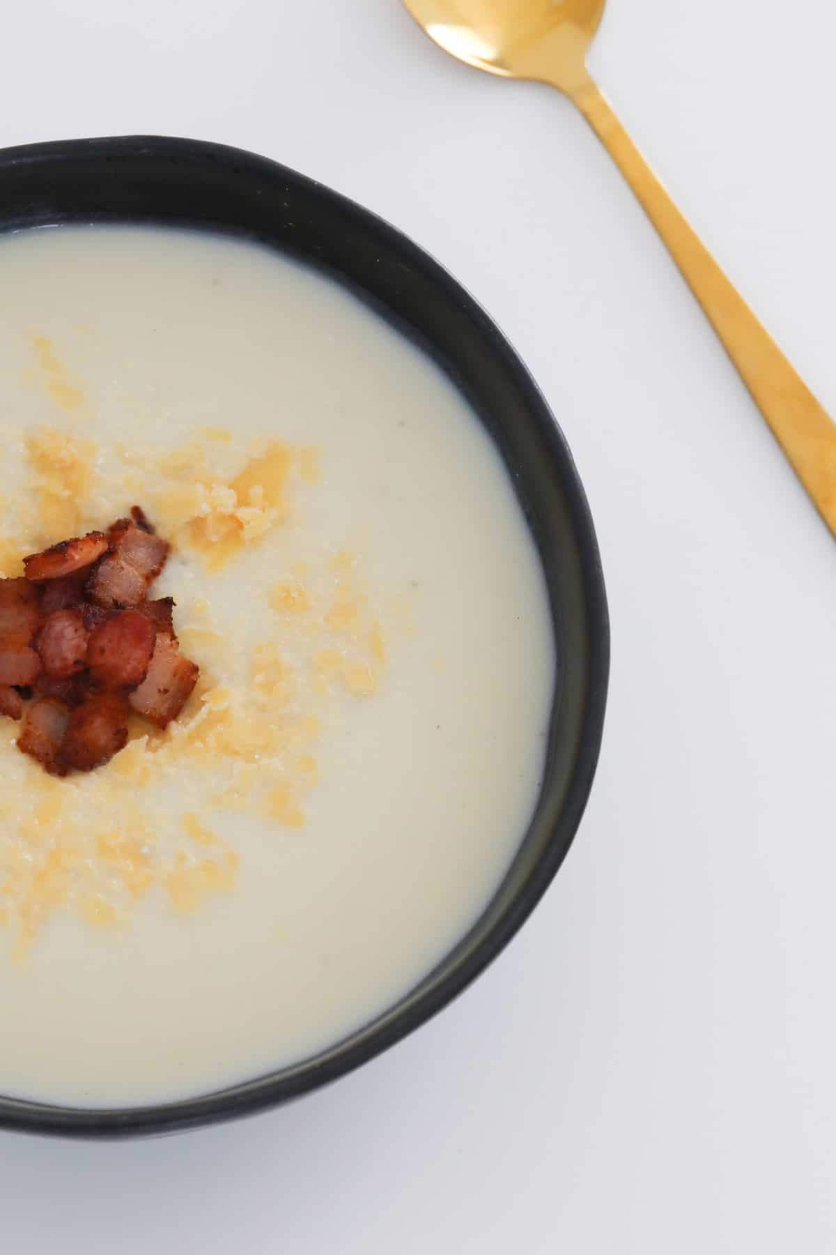 A black bowl with creamy soup garnished with grated parmesan and bacon pieces with a gold spoon beside it