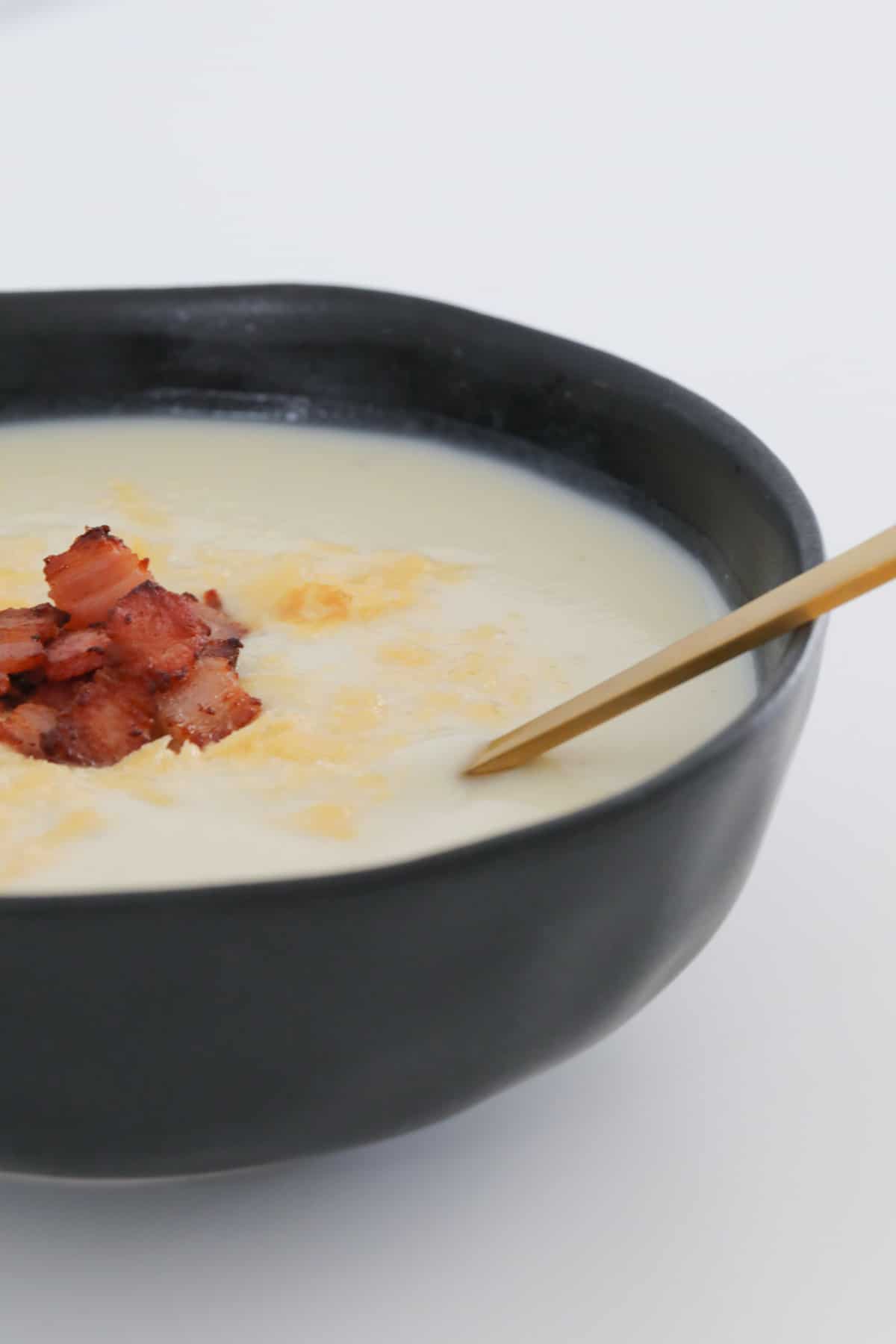 A close up image of a golden spoon resting in a bowl of white soup with bacon and parmesan cheese garnish