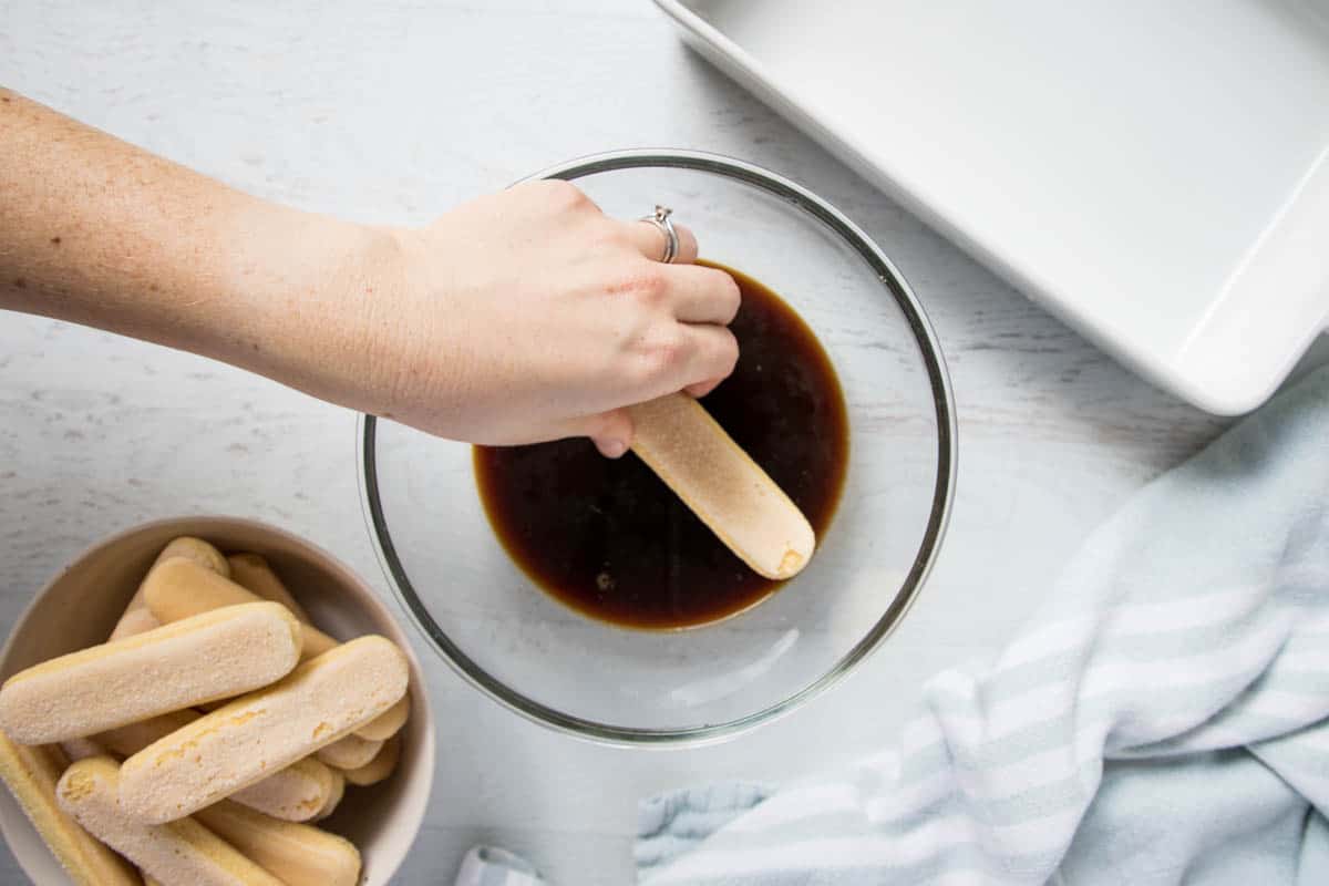 a hand dipping a ladyfinger sponge in coffee mixture