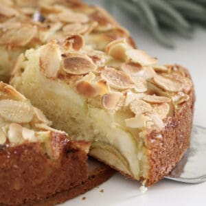 A slice of butter cake with layeres of apples and almonds on top being removed from the cake.