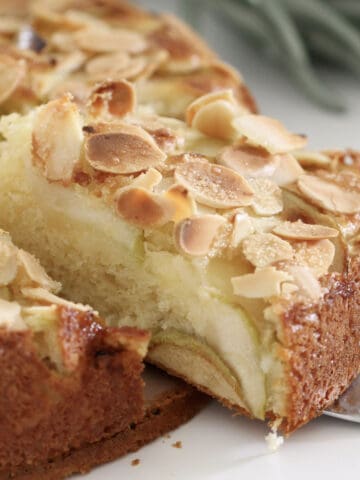 A slice of butter cake with layeres of apples and almonds on top being removed from the cake.