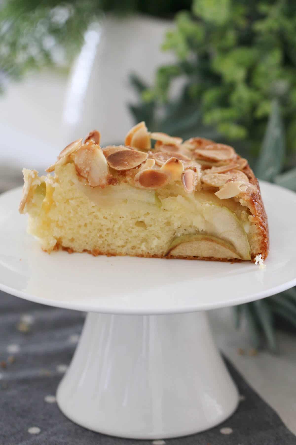 A slice of a layered apple and almond cake on a white cake stand.