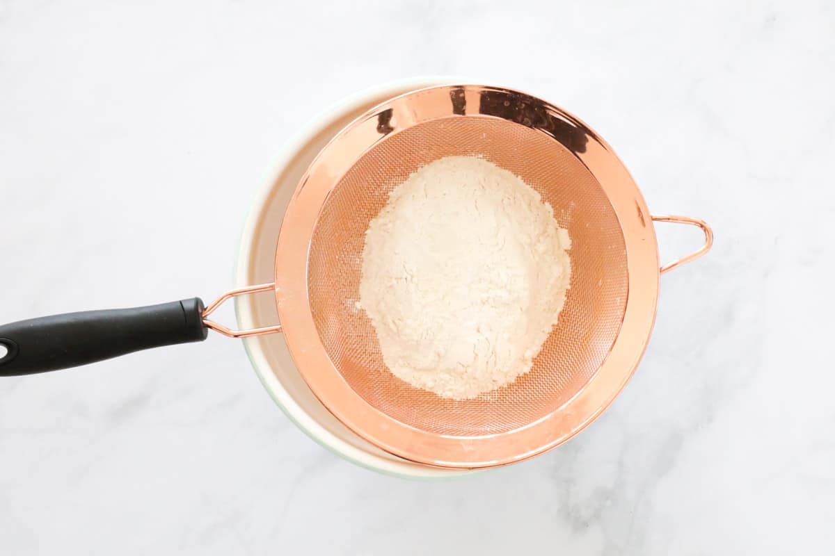 Flour being sifted into a bowl using a copper sieve.