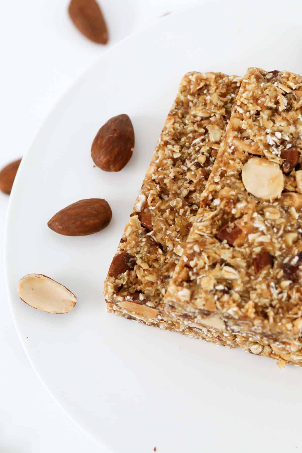 Almond, oat and date muesli bars on a white cake stand with roasted almonds scattered nearby.