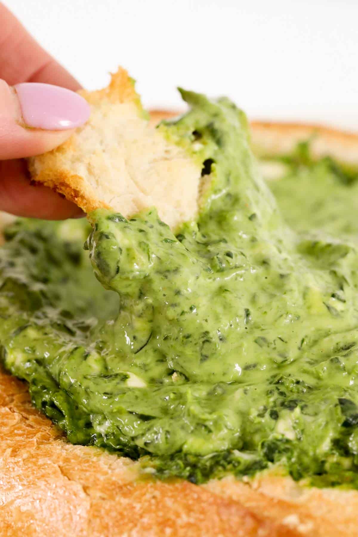 A hand dipping a piece of bread into green spinach dip in a cob loaf.
